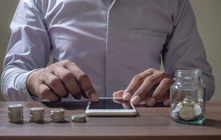 Ein Mann, der ein Handy auf einem Schreibtisch bedient, während Münzen in einem Glas und auf einem Tisch gestapelt liegen