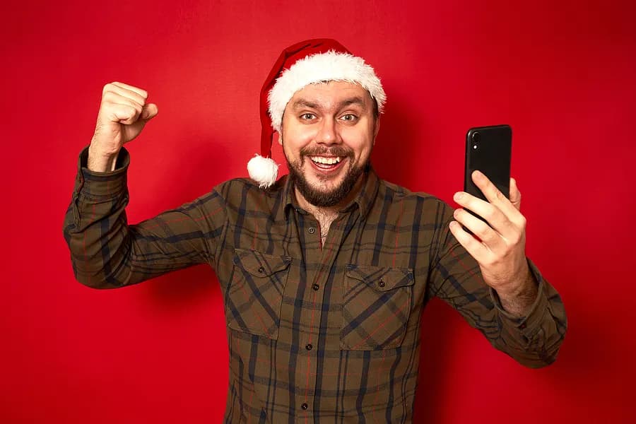 Ein erwachsener Mann mit einer Weihnachtsmannmütze, der begeistert ein Handy in der Hand hält