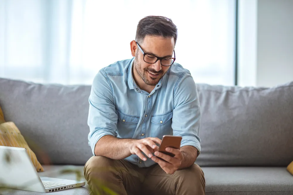 Ein Mann mit Brille und blauem Hemd sitzt auf einem grauen Sofa und lächelt in sein Handy, das er in den Händen hält.