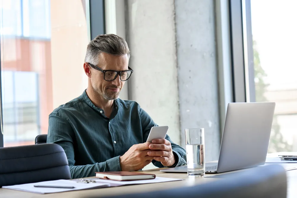Mann, der während der Arbeit am Laptop auf sein Smartphone schaut