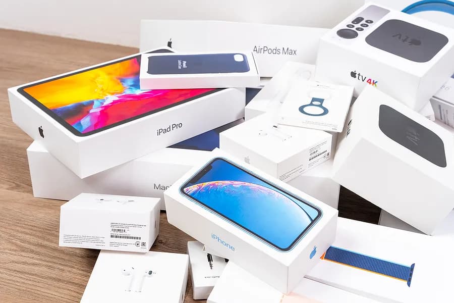 Ein Stapel von Apple-Produktverpackungen, darunter iPhone, iPad Pro, AirPods, Apple Watch und Apple TV, auf einer Holzfläche angeordnet.