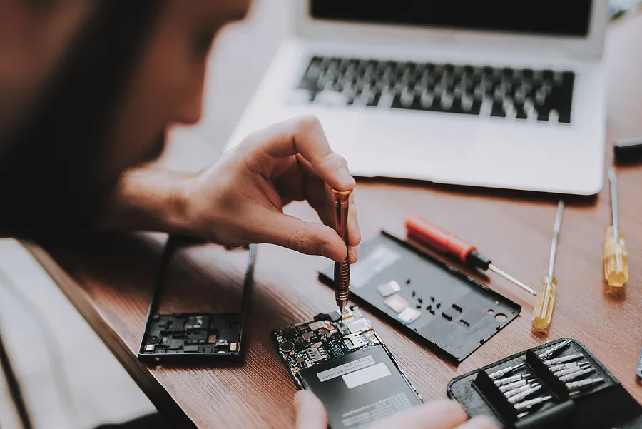 Ein Techniker repariert mit einem Präzisionsschraubendreher die inneren Bauteile eines Smartphones auf einem Holztisch, umgeben von Kleinwerkzeug, Ersatzteilen und einem geöffneten Laptop.