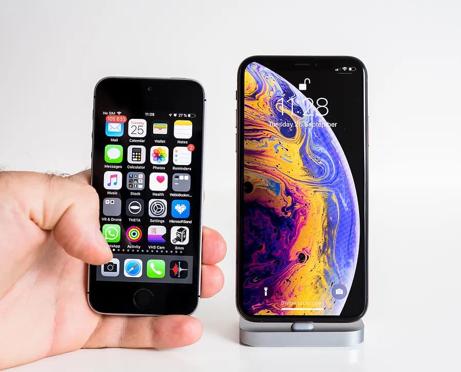 Eine Hand, die ein kleines, älteres iPhone neben einem größeren, modernen iPhone auf einem Ständer hält, um die Unterschiede in Größe und Design zwischen einem alten iPhone und einem neueren Modell zu verdeutlichen.
