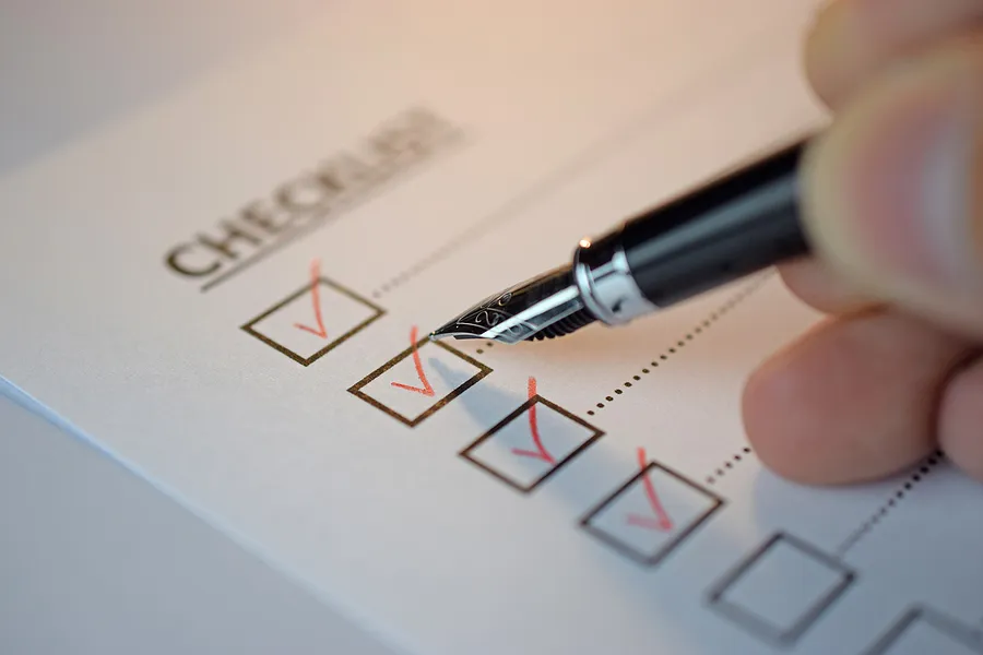 Nahaufnahme einer Hand, die mit einem Stift Punkte auf einer Checkliste abhakt; dargestellt wird eine alte Checkliste zur Prüfung eines Telefons hinsichtlich Akku, Leistung und Sicherheit.